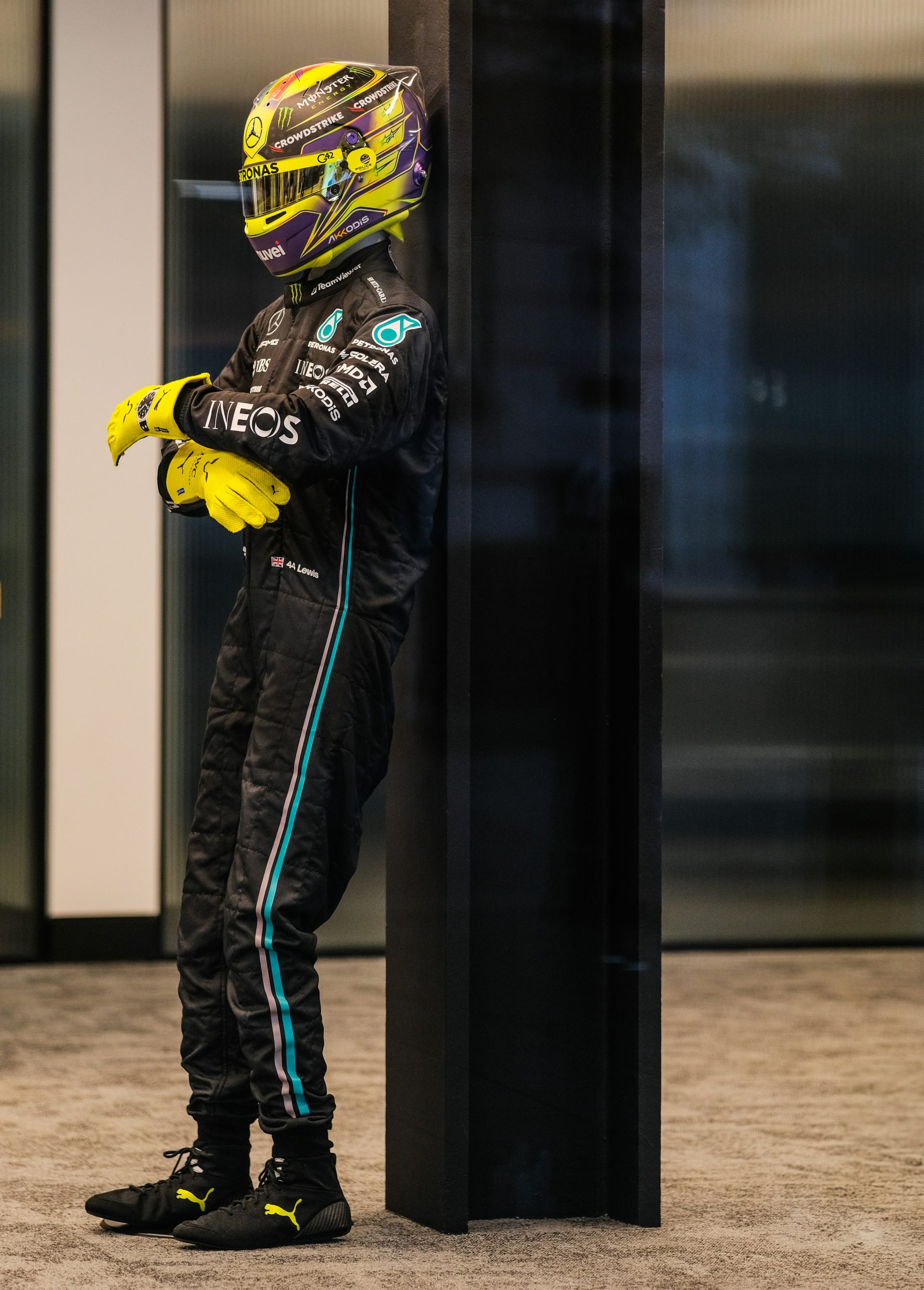 Formule‑1‑coureur in zwart‑turquoise racepak met helm en gekruiste armen, leunend tegen een donkere wand.