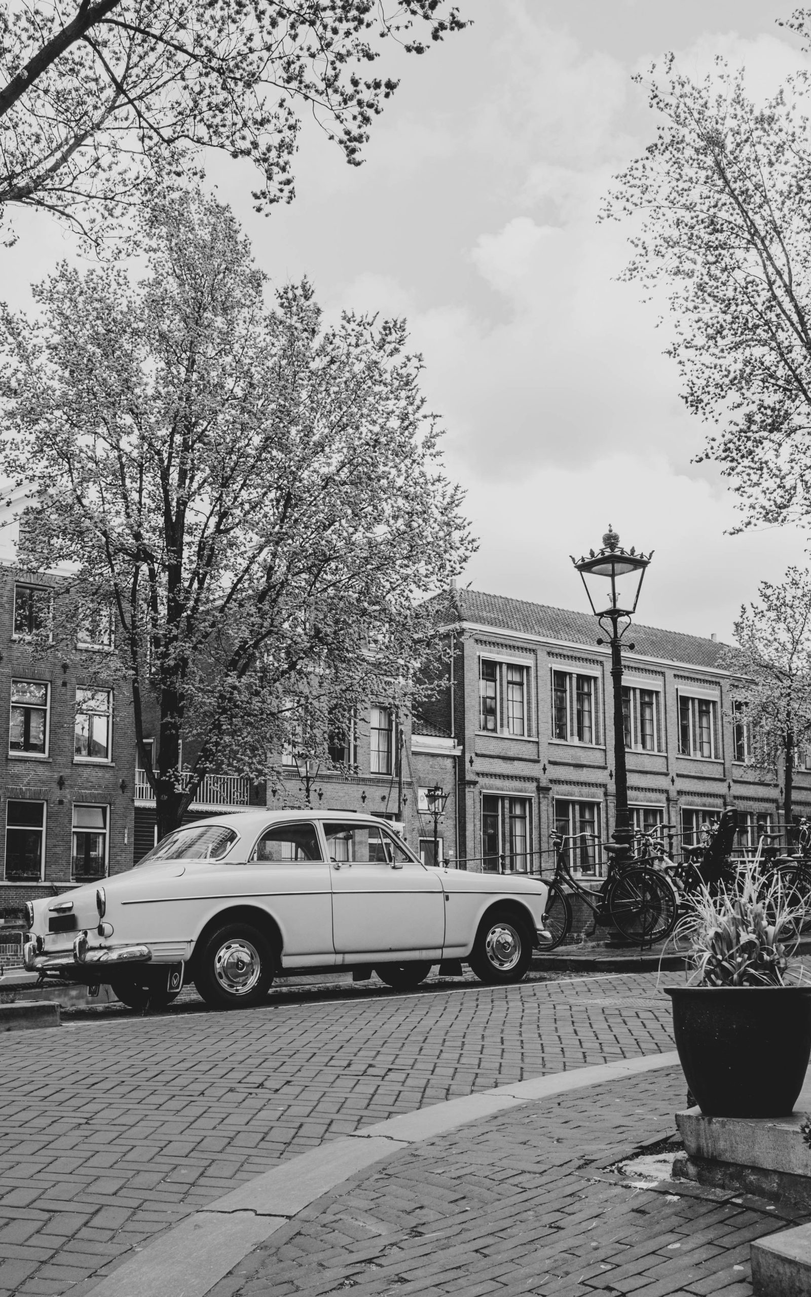Zwart‑wit straatbeeld met een klassieke auto op een kasseienweg, omringd door fietsen, bomen en oude Europese gebouwen.