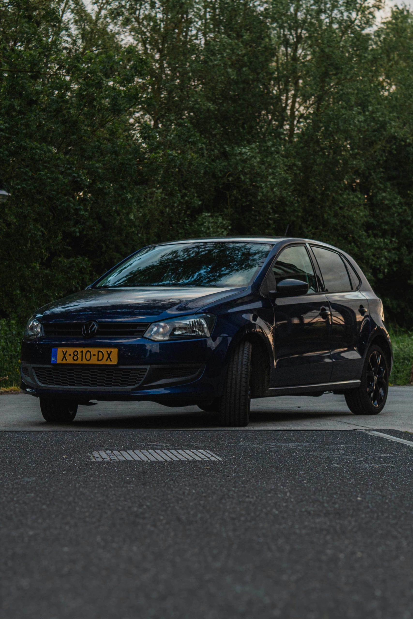 Donkerblauwe hatchback (Volkswagen Polo) geparkeerd op een parkeerplaats met bomen op de achtergrond in de schemering.