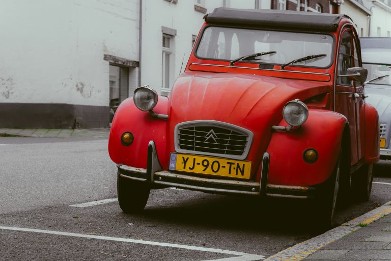 Rode Citroën 2CV met zwart dak geparkeerd in een Europese straat met bakstenen gevels.