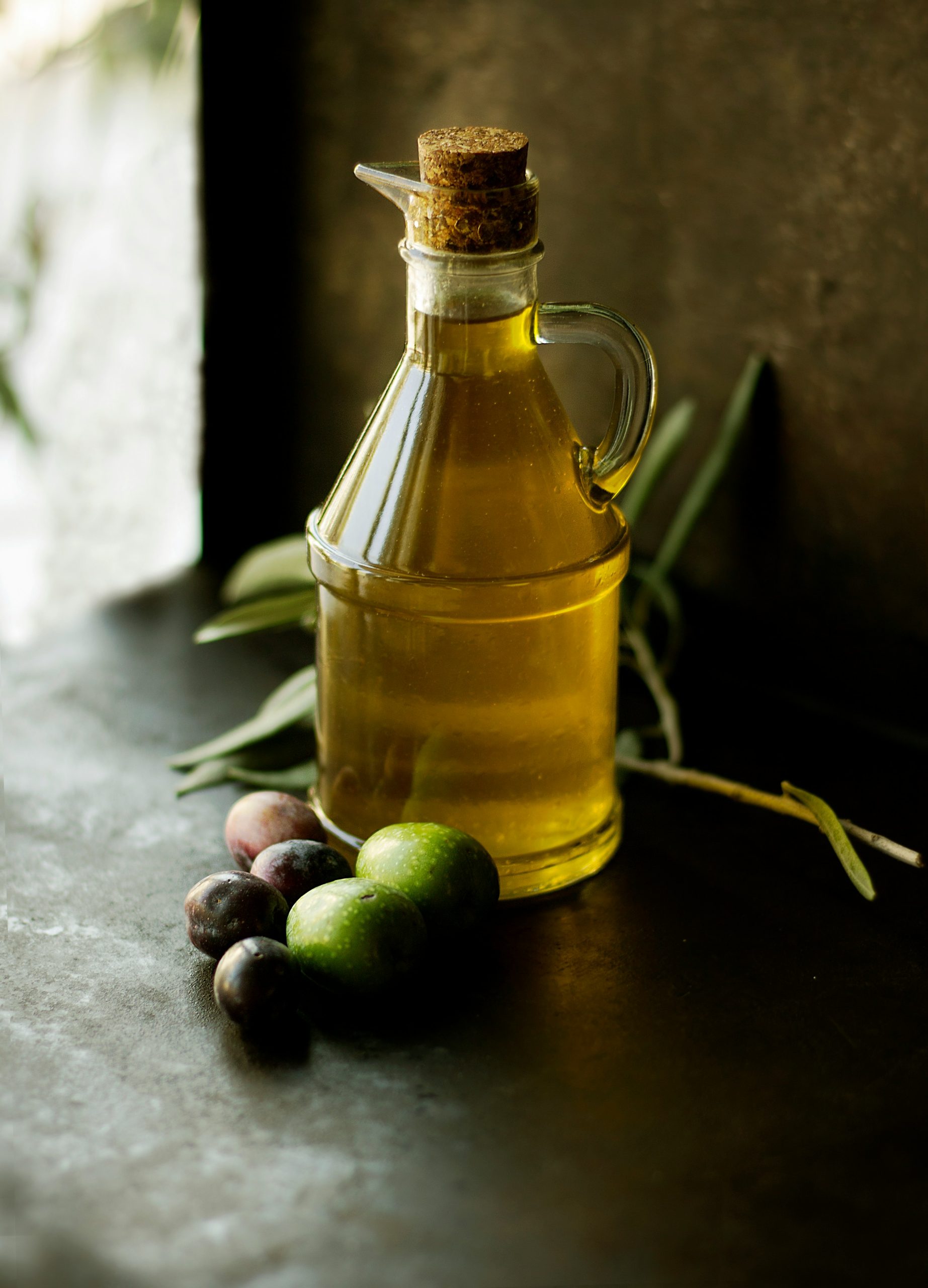 Glazen fles met goudgele olie naast groene en paarse olijven en olijfbladeren op een donkere ondergrond.
