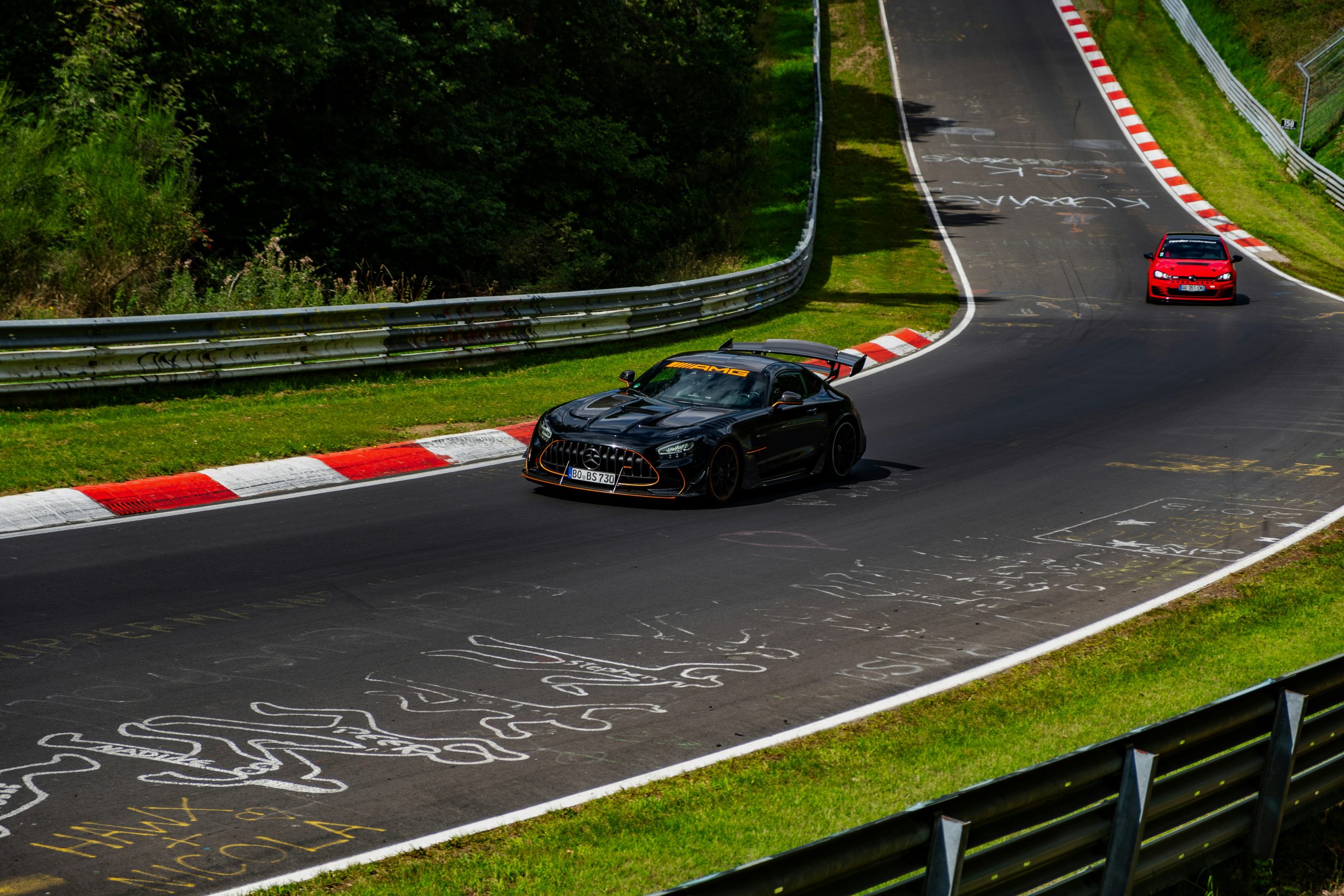 Zwarte Mercedes‑AMG GT Black Series met oranje accenten op een circuit, gevolgd door een rode auto, met kleurrijke markeringen op het asfalt.