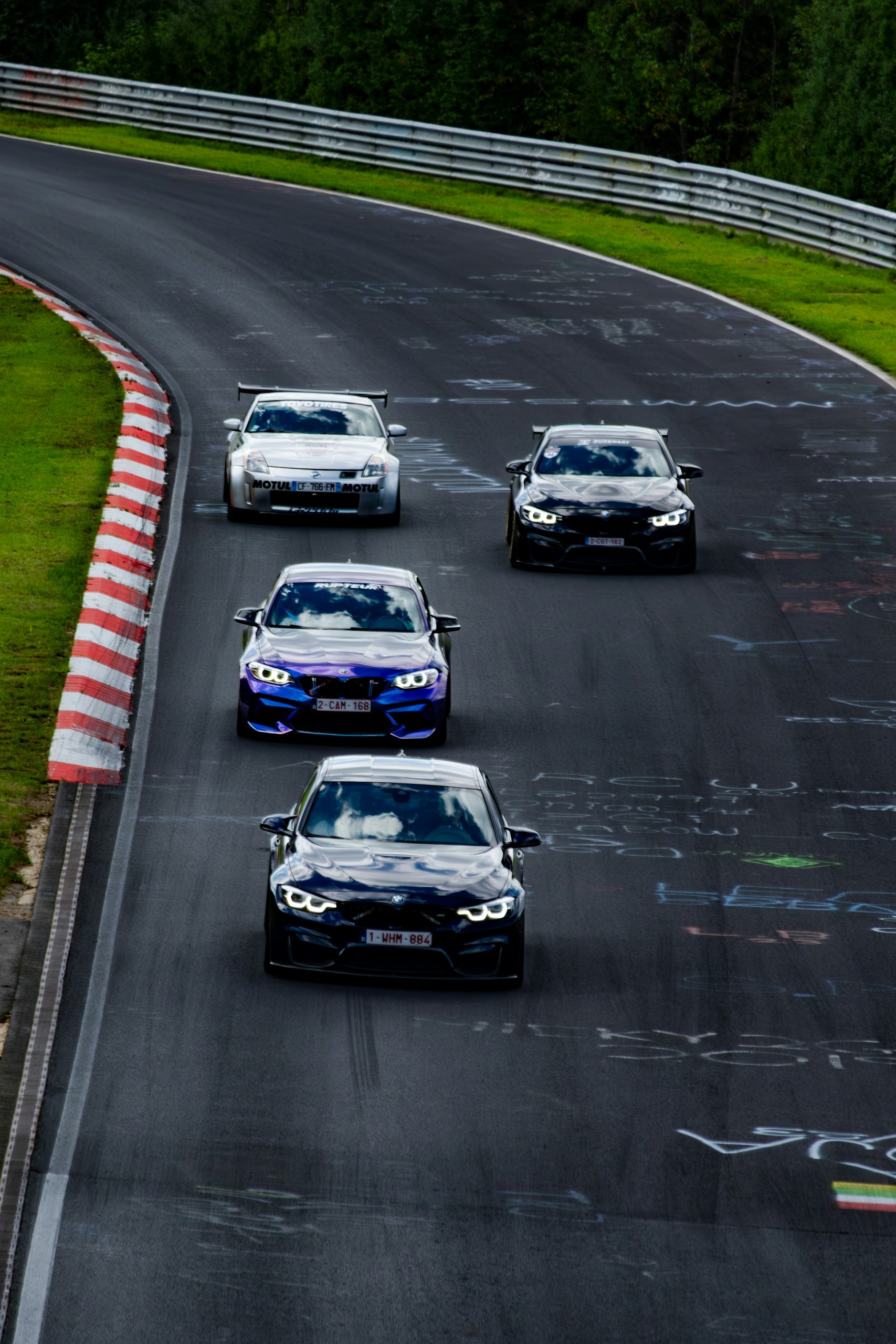 Vier sportwagens dicht op elkaar op een circuit met rood‑witte curbstones en graffiti op het asfalt, omringd door groen en vangrails.