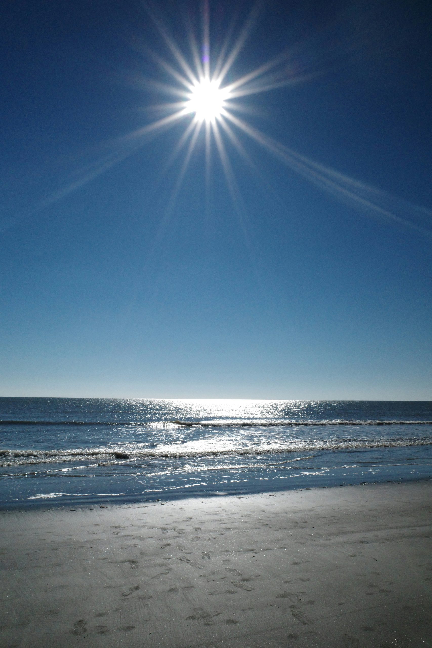 Felle zon boven een rustige zee met lichtreflecties op het water en voetstappen in het natte strandzand.
