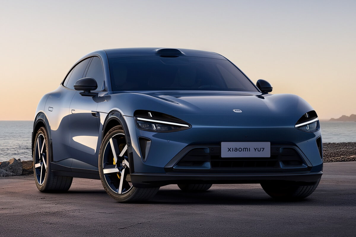Zilveren Xiaomi‑elektrische auto geparkeerd aan een kustweg bij zonsondergang, met zee en rotsformaties op de achtergrond.