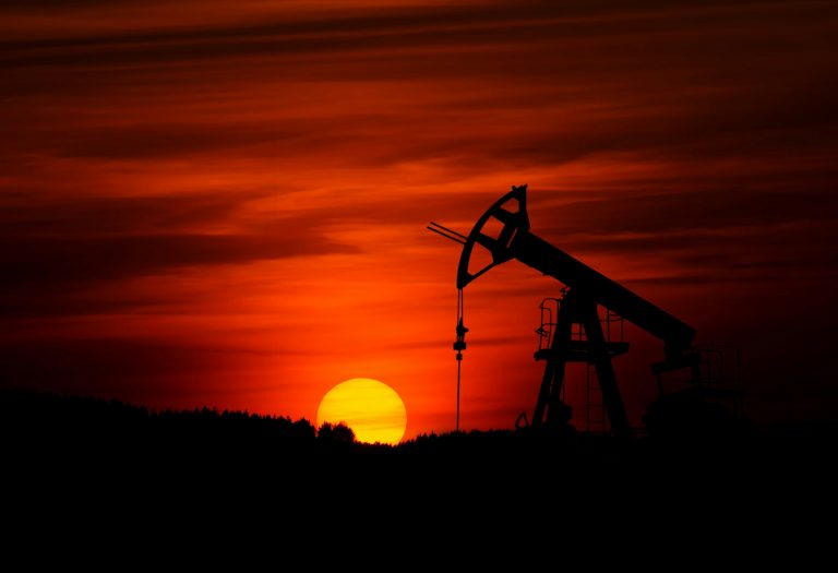 Silhouet van een olie‑pompinstallatie tegen een felrode zonsondergang met bomen op de horizon.