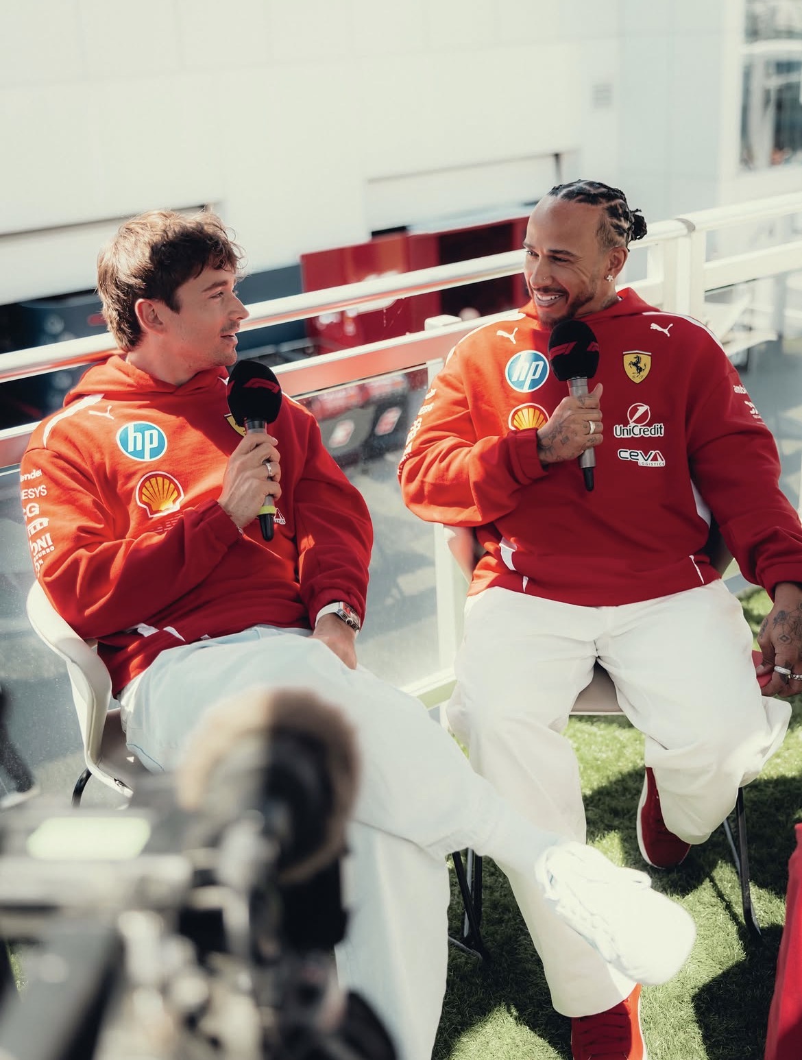 Twee personen in rode Ferrari‑teamkleding die buiten op stoelen zitten en praten met microfoons, met een camera op de voorgrond.