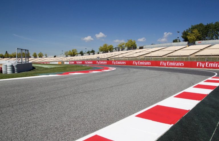 Asfaltcircuit met rood‑witte kerbstones, bandenstapels en tribunes onder een heldere blauwe lucht.