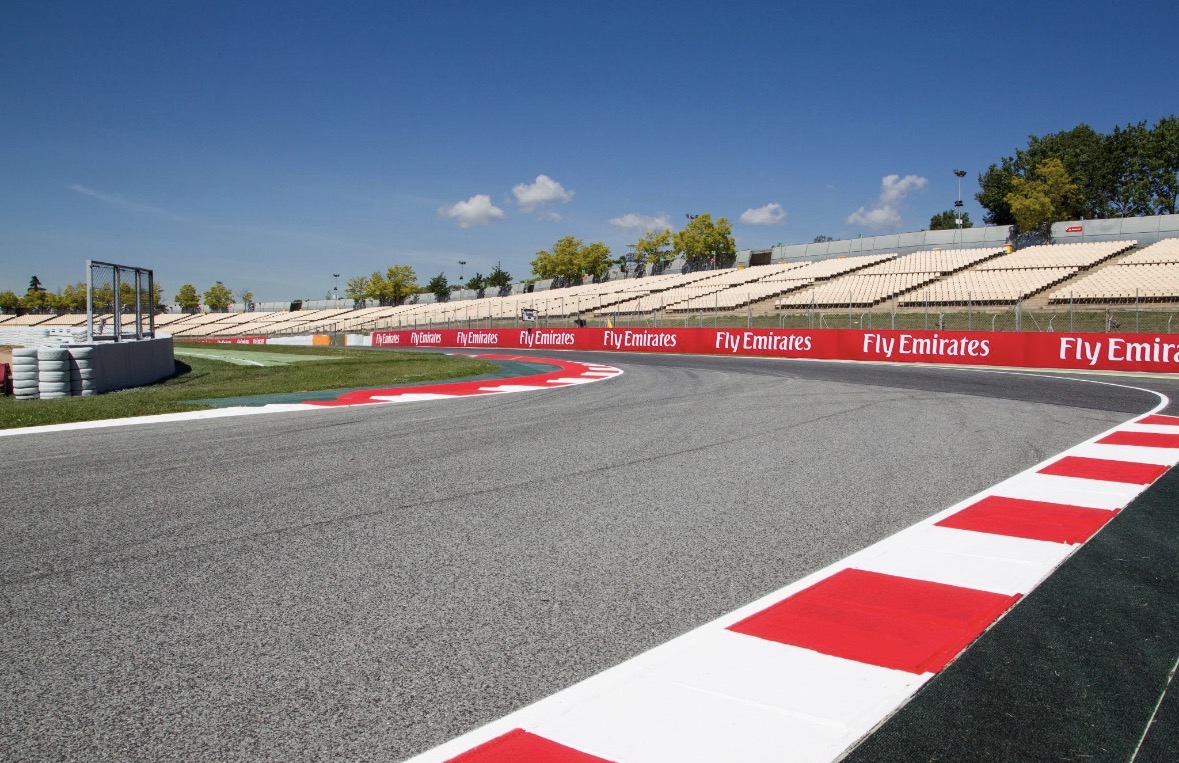 Asfaltcircuit met rood‑witte kerbstones, bandenstapels en tribunes onder een heldere blauwe lucht.