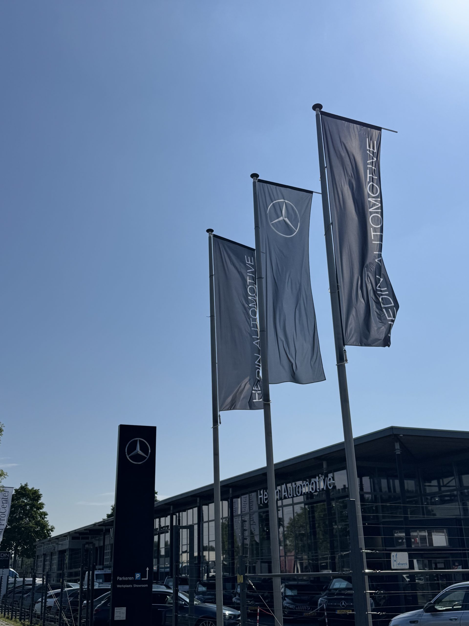 Drie vlaggen met het Mercedes-logo wapperen voor een modern Mercedes-Benz autodealer gebouw.