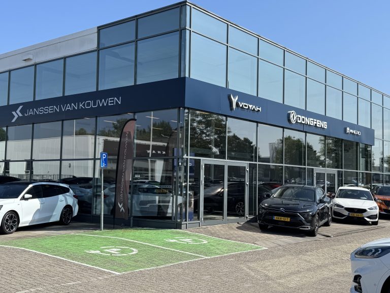 Janssen van Kouwen-dealership met Voyah en Dongfeng-logo’s, moderne glazen gevel, en enkele geparkeerde auto’s inclusief een groene laadplek.