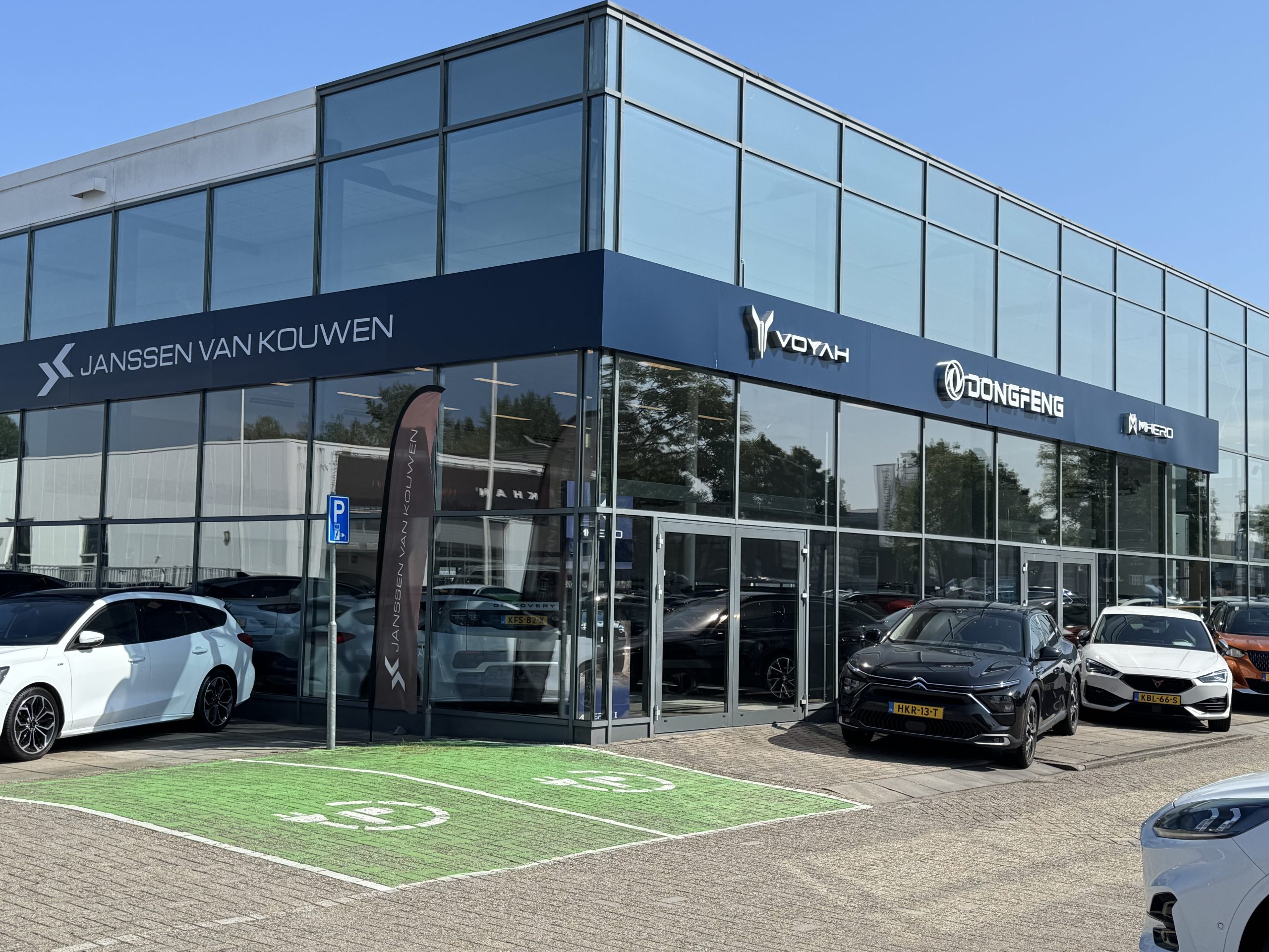 Janssen van Kouwen-dealership met Voyah en Dongfeng-logo’s, moderne glazen gevel, en enkele geparkeerde auto’s inclusief een groene laadplek.