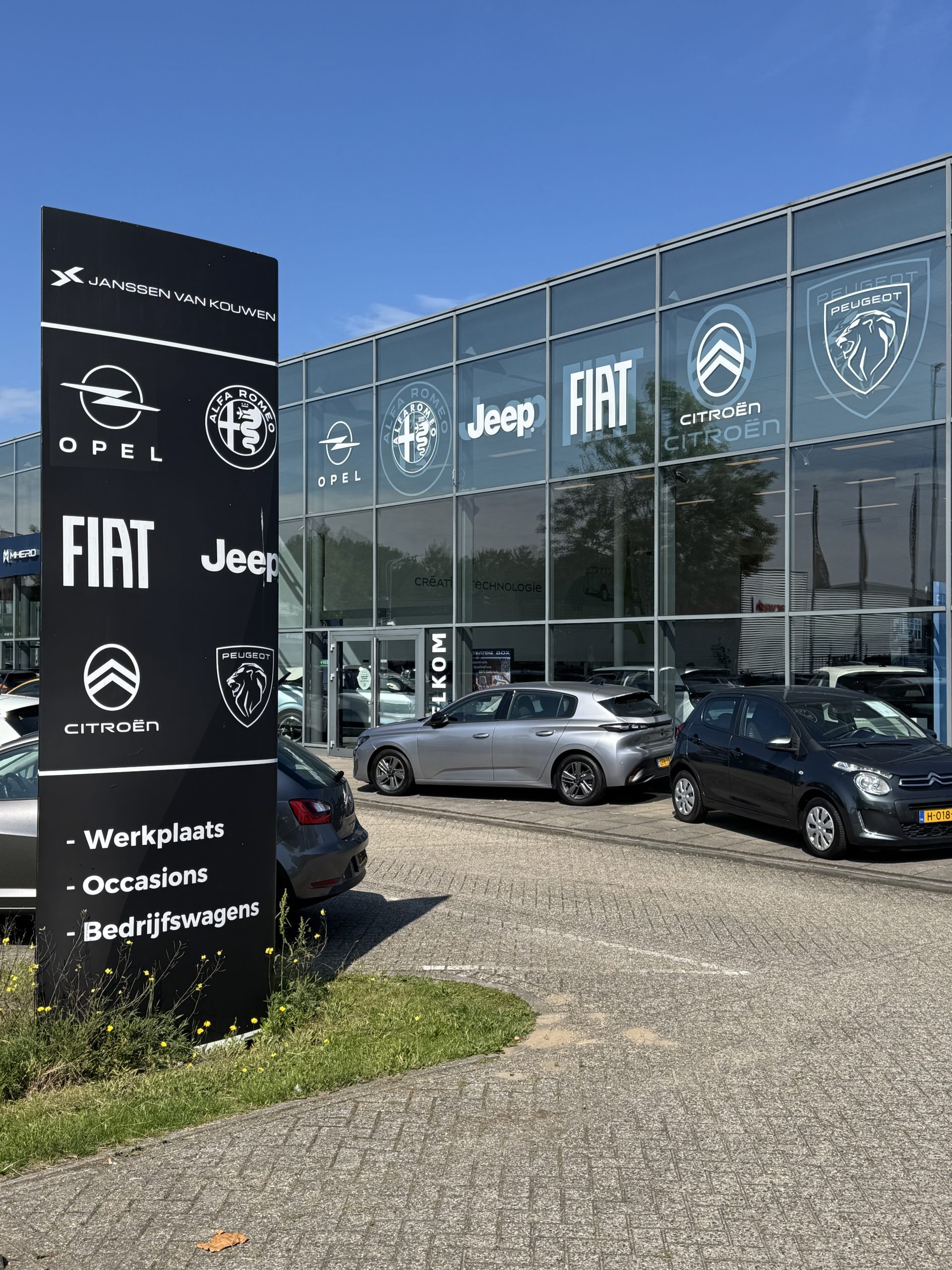 Janssen van Kouwen-dealership met groot bord waarop merken als Opel, Fiat, Jeep, Citroën en Peugeot staan, met auto’s voor het glazen gebouw.