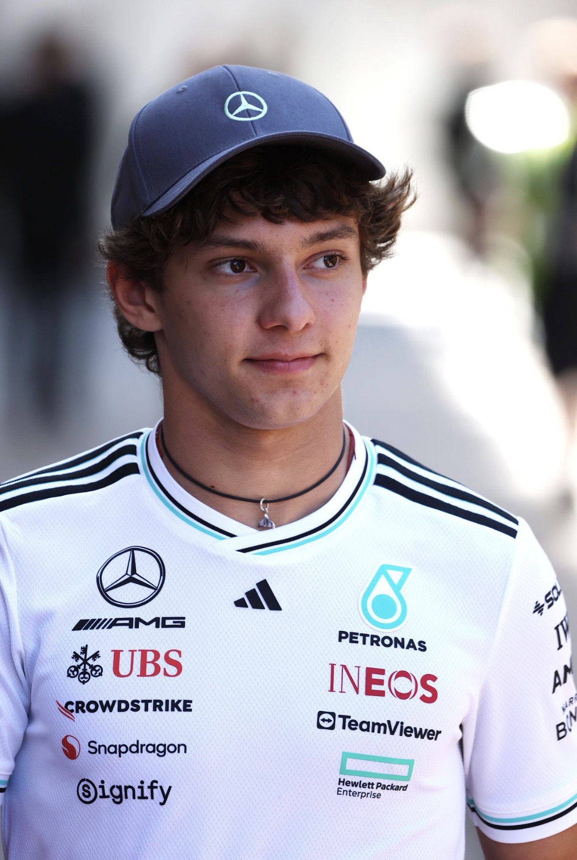 Persoon met grijze Mercedes‑pet en witte teamshirt vol sponsoren, gefotografeerd buiten met wazige achtergrond.