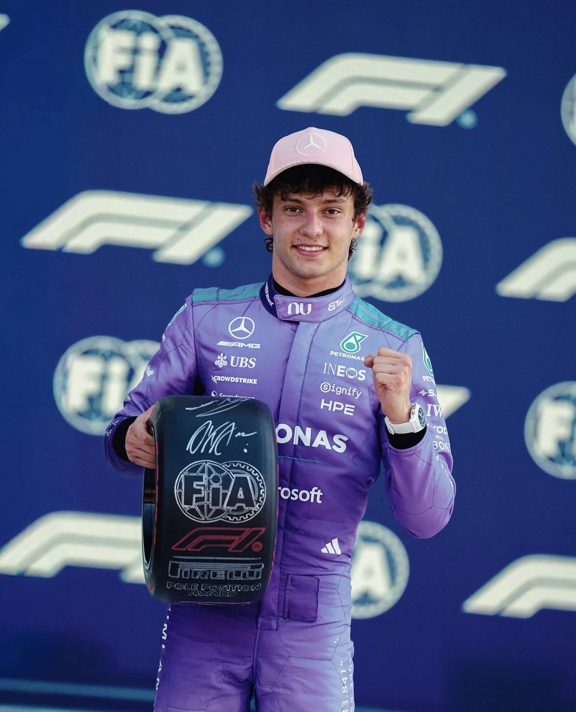 Formule‑1‑coureur in paarse teamkleding met roze pet, poserend met een zwarte Pirelli pole‑award voor een FIA‑achterwand.