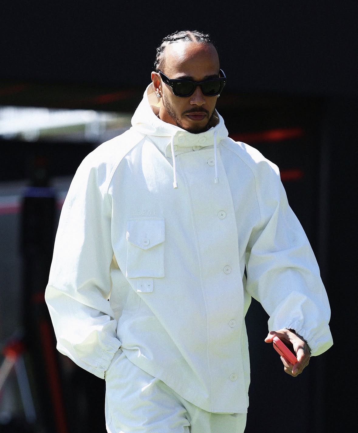 Persoon in oversized witte jas en broek met zonnebril en rode telefoon, staand voor een donkere buitenlocatie.