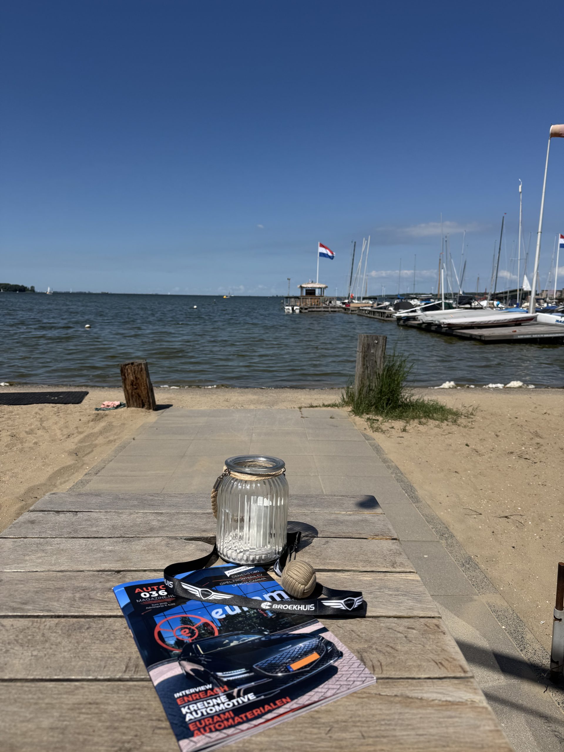 Zonnige waterfront‑setting met AUTO036‑magazine, Broekhuis‑lanyard en nautische decoratie bij een jachthaven.