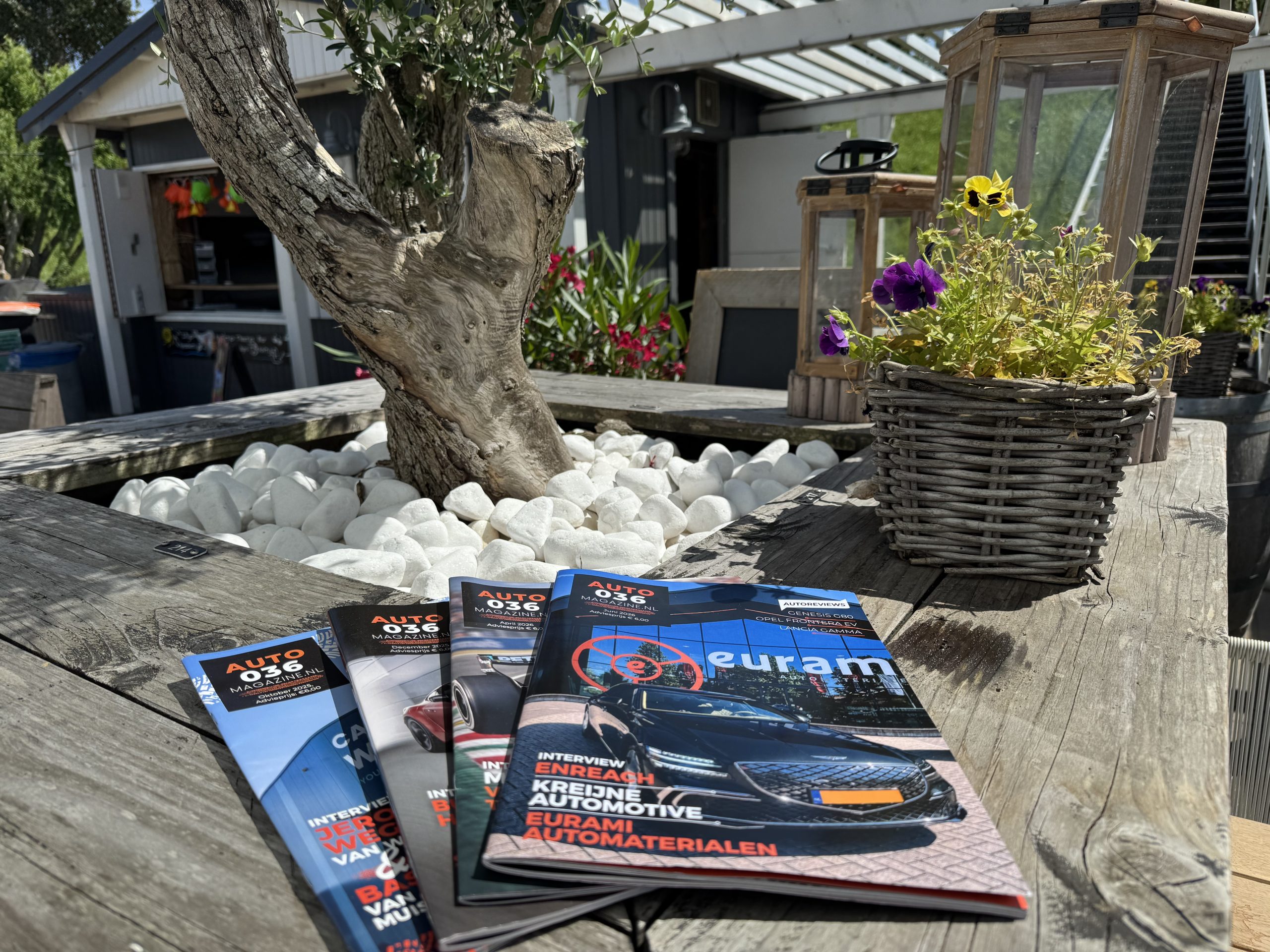 Drie AUTO036‑magazines op een houten buitentafel met bloemen en decoratieve lantaarns bij een terrasomgeving.