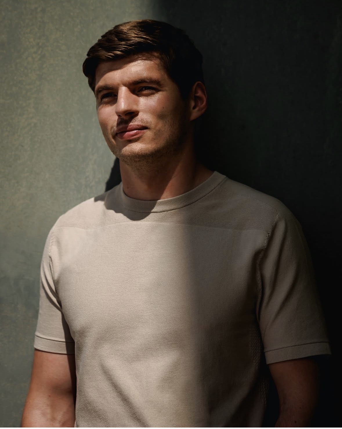 Persoon in beige T‑shirt staand tegen een muur, deels in de schaduw met sterk contrast door zonlicht.