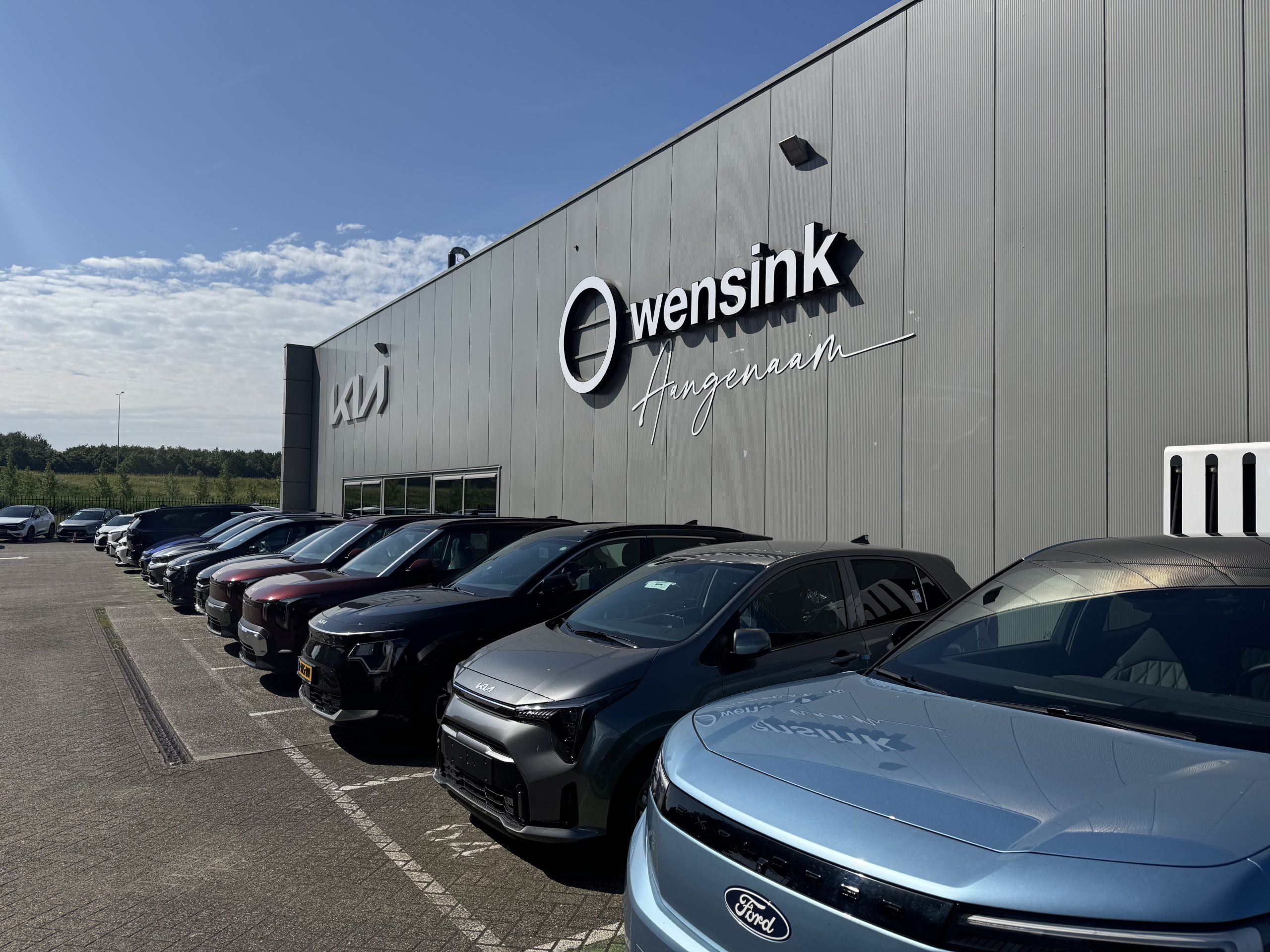 Kia-dealership Wensink Angenaam met rij nieuwe auto’s voor het gebouw onder een heldere blauwe lucht.