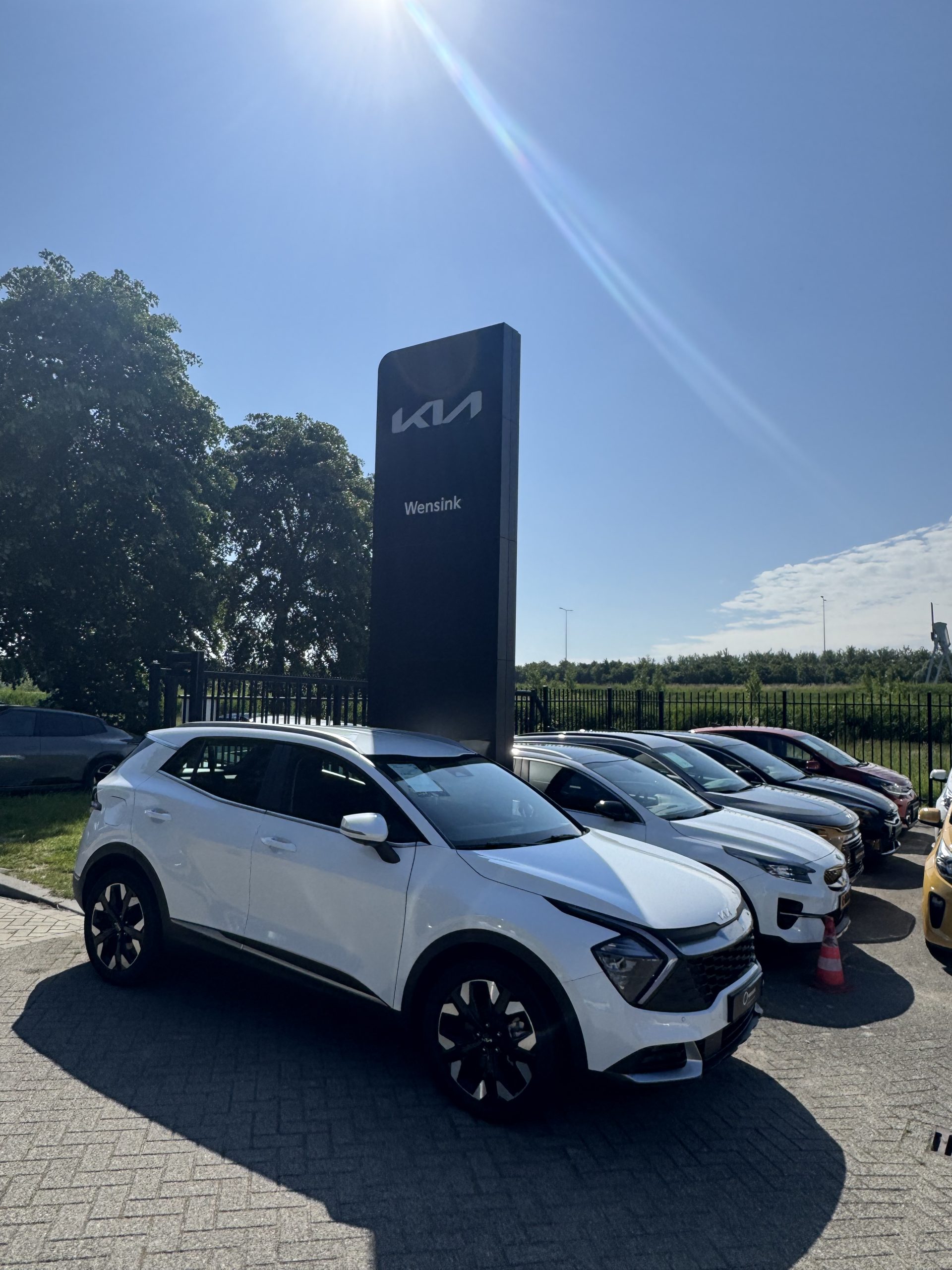 Witte Kia SUV geparkeerd voor een groot Kia-bord bij Wensink Angenaam, met meer auto’s op de achtergrond.