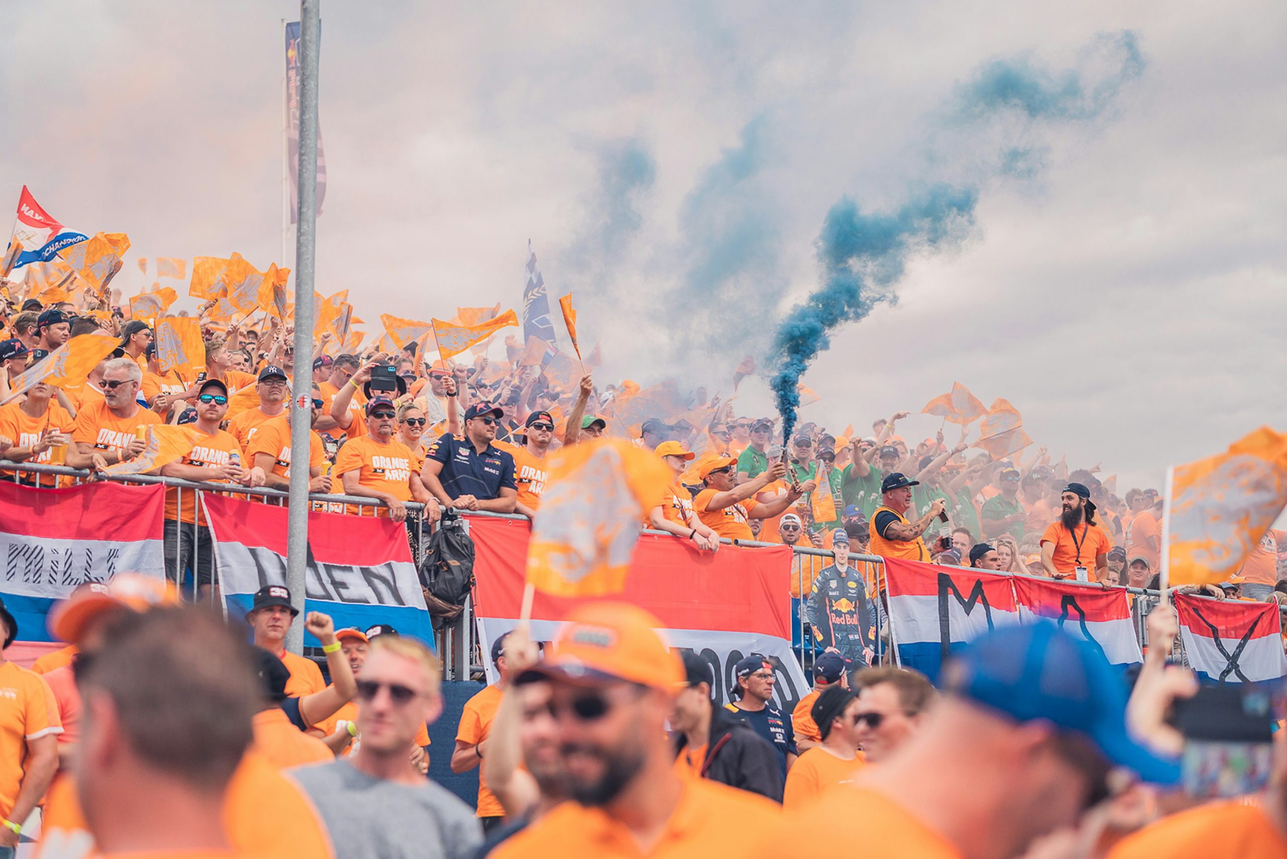 Grote groep fans in oranje shirts en vlaggen juichend op een tribune, met blauwe rook en Nederlandse vlaggen.