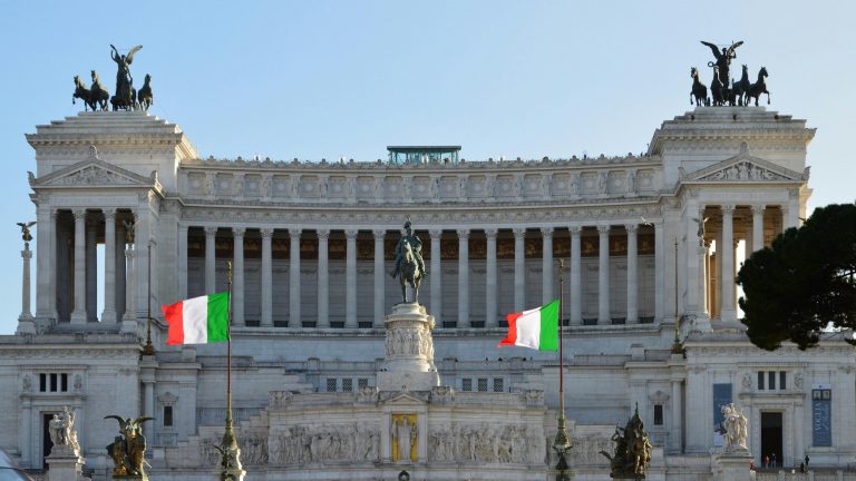 Groots wit marmeren monument met ruiterstandbeeld, zuilen en twee Italiaanse vlaggen aan de voorzijde.
