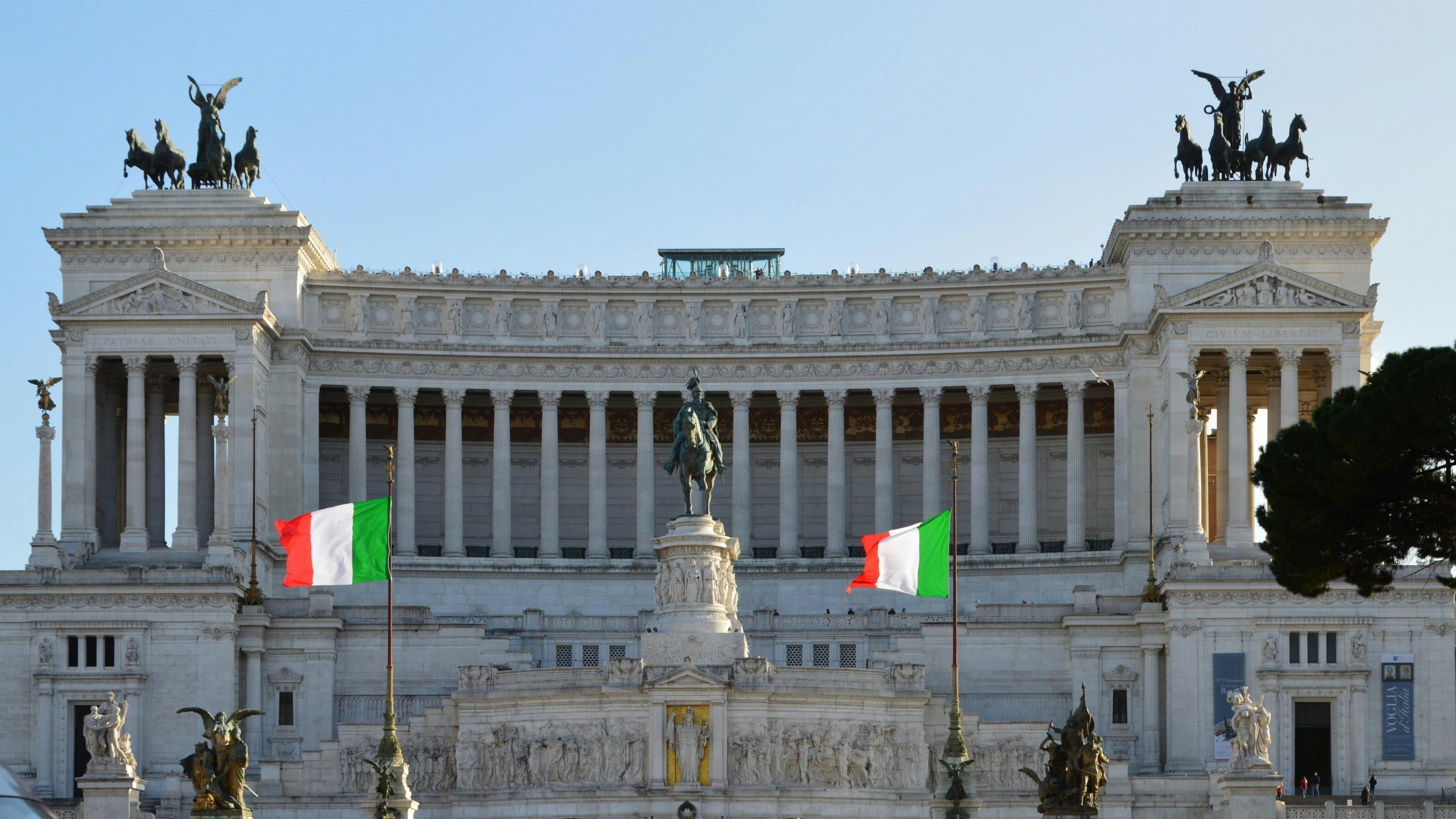 Groots wit marmeren monument met ruiterstandbeeld, zuilen en twee Italiaanse vlaggen aan de voorzijde.