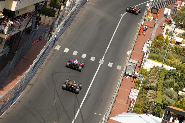 Formule 1-auto's van Ferrari en McLaren razen over het smalle stratencircuit van Monaco, terwijl fans vanaf balkons boven de vangrails naar beneden leunen.