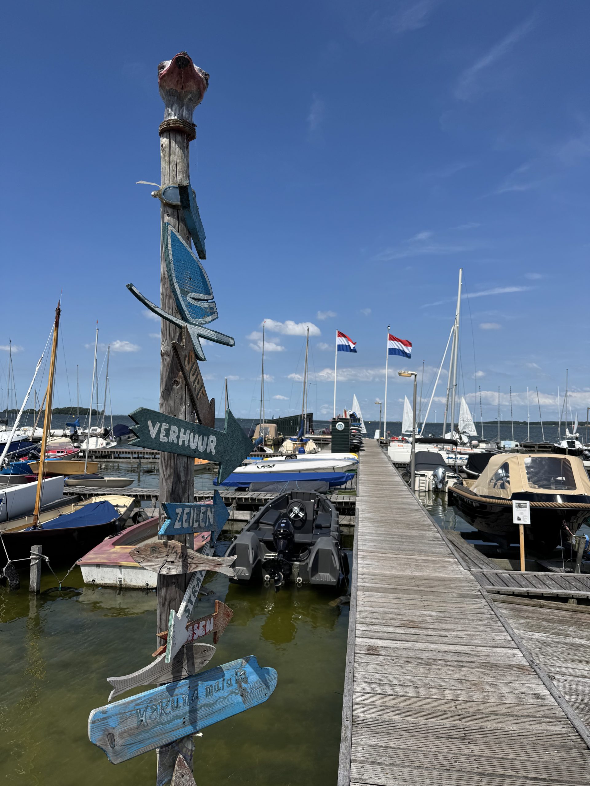 Jachthaven met houten wegwijzers voor verhuur, zeilen en horeca langs een steiger met meerdere boten.