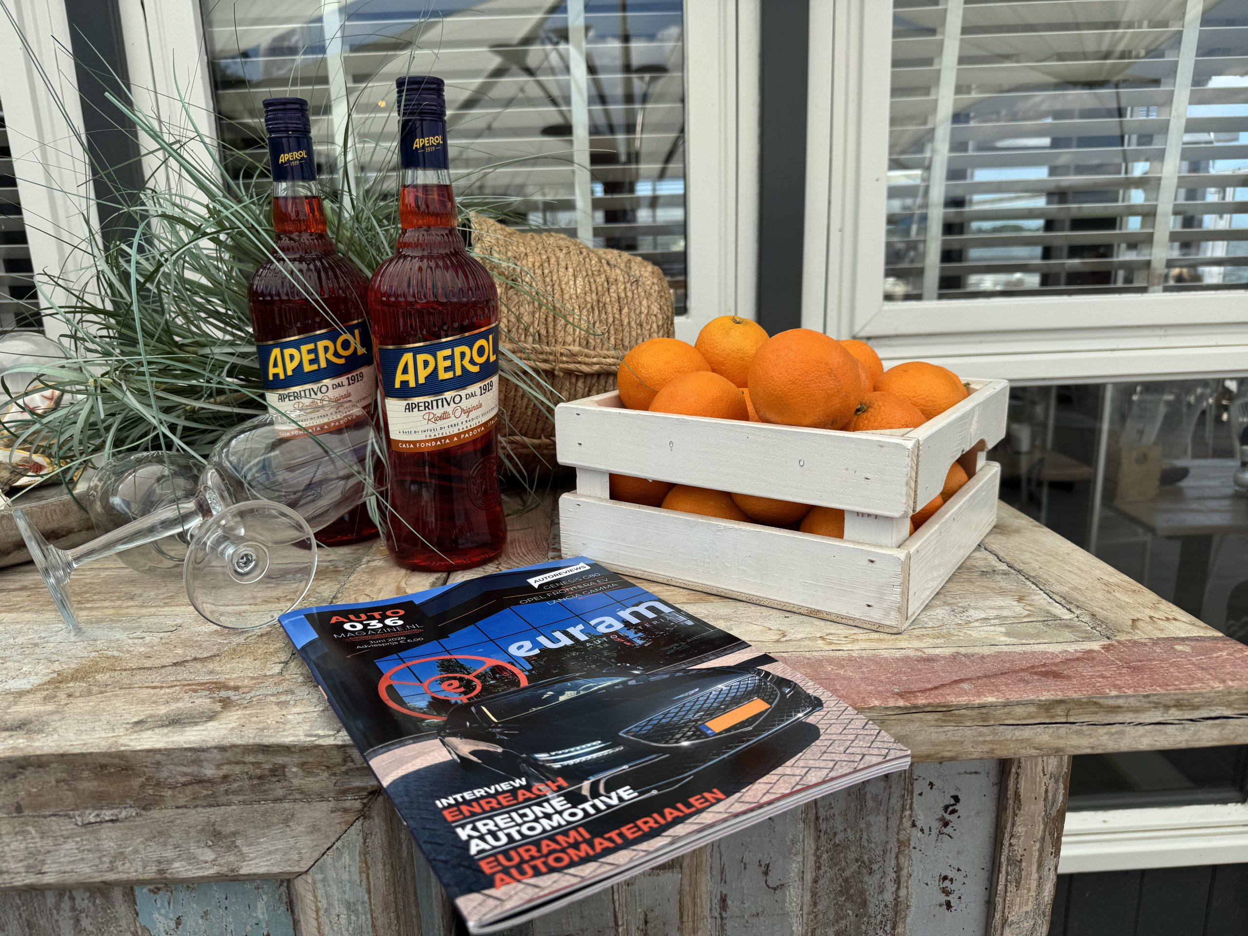 Rustieke tafel met Aperol‑flessen, verse sinaasappels en een automotive magazine in een zonnige patio‑omgeving.