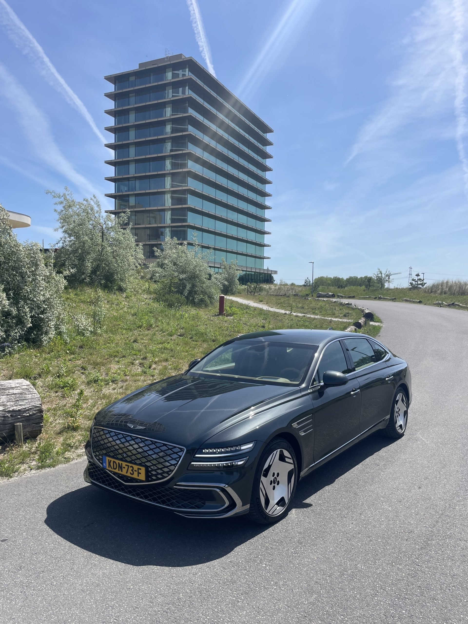 Zwarte sedan met kenteken KDN‑73‑F geparkeerd naast modern glazen kantoorgebouw onder blauwe lucht met contrails.