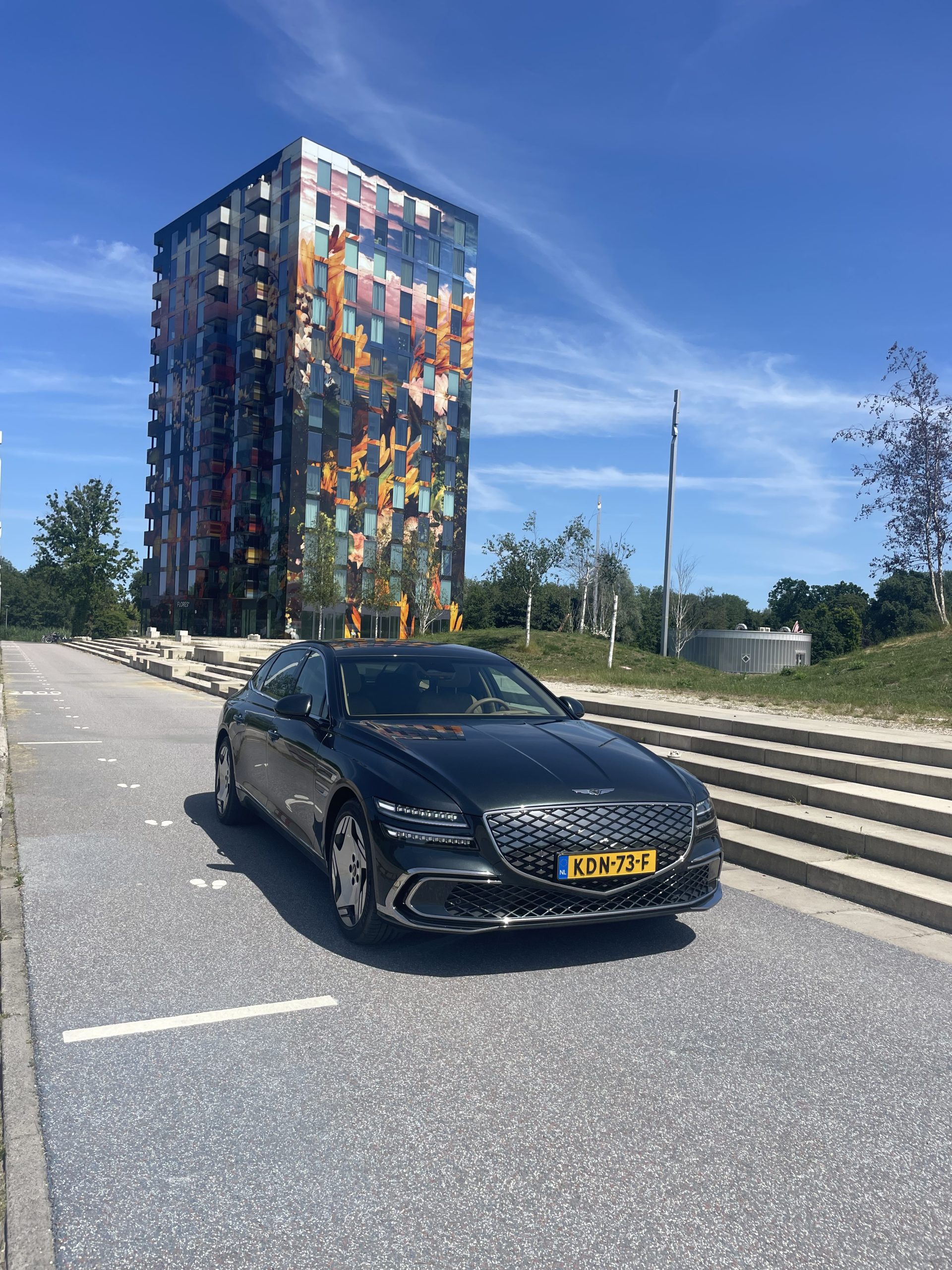 Moderne zwarte sedan met kenteken KDV‑73‑F geparkeerd voor een gebouw met opvallende kleurrijke muurschildering.