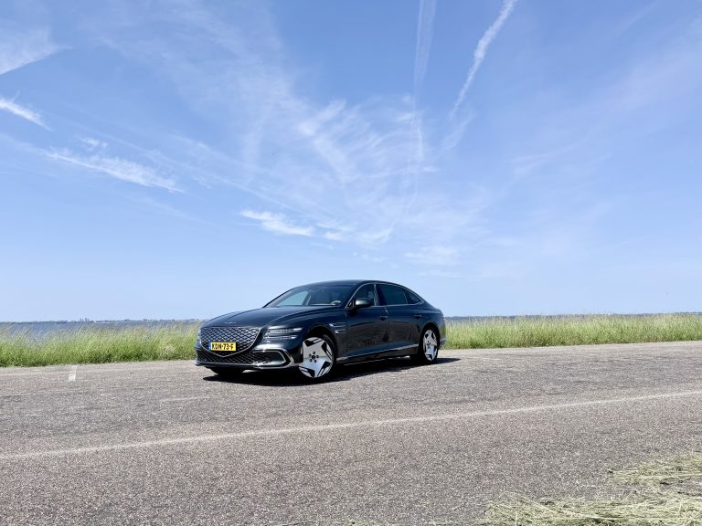 Zwarte luxe sedan met kenteken KDN‑73‑F geparkeerd langs een rustige weg bij water en gras onder heldere lucht.