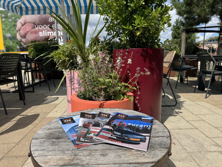 Buitenterras met houten tafel, automotive magazines en grote plantenpotten onder blauw‑wit zonnescherm.