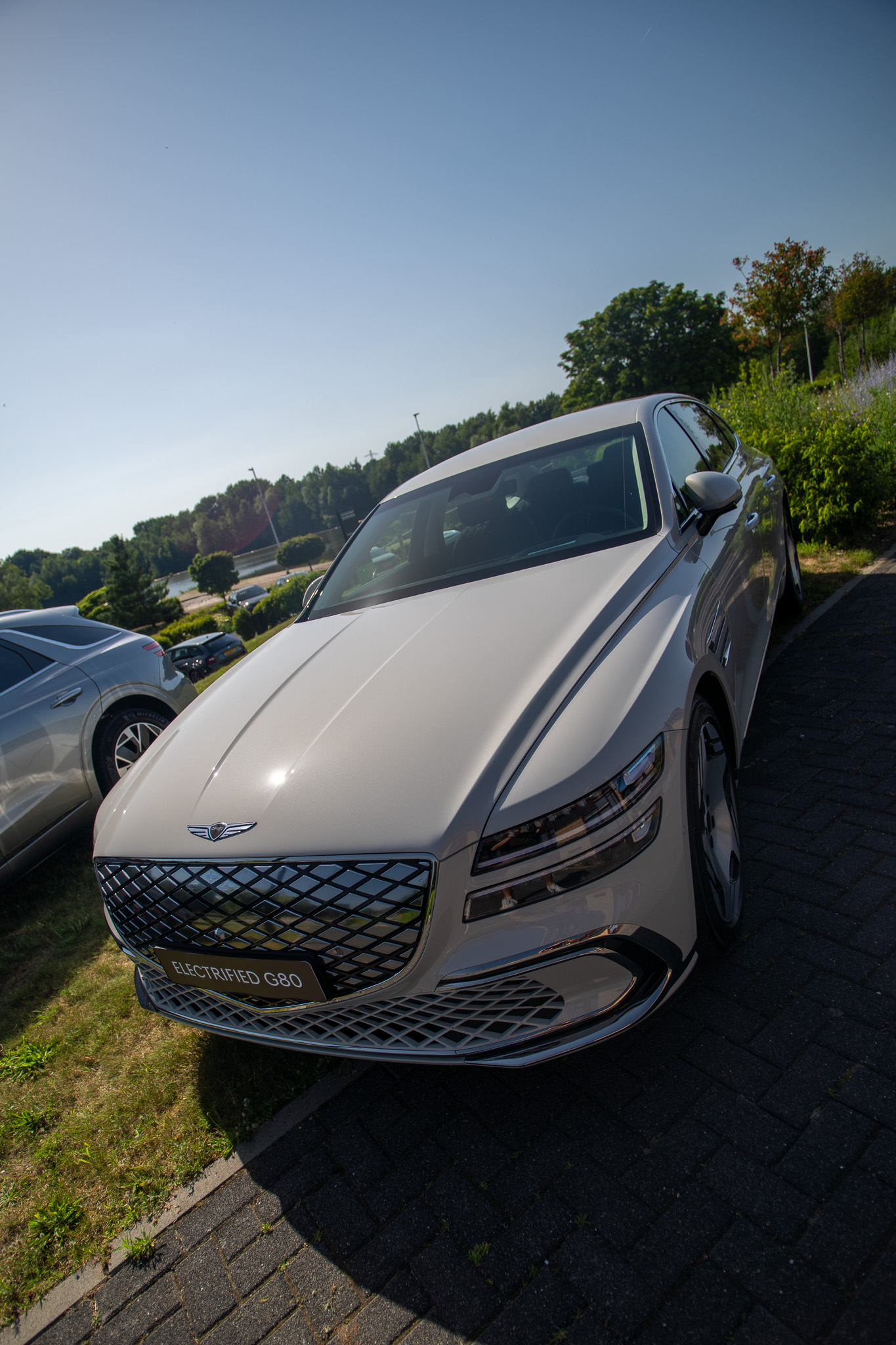 Witte Genesis Electrified G80 met diamantgrille geparkeerd in zonnige omgeving met water en bomen op de achtergrond.