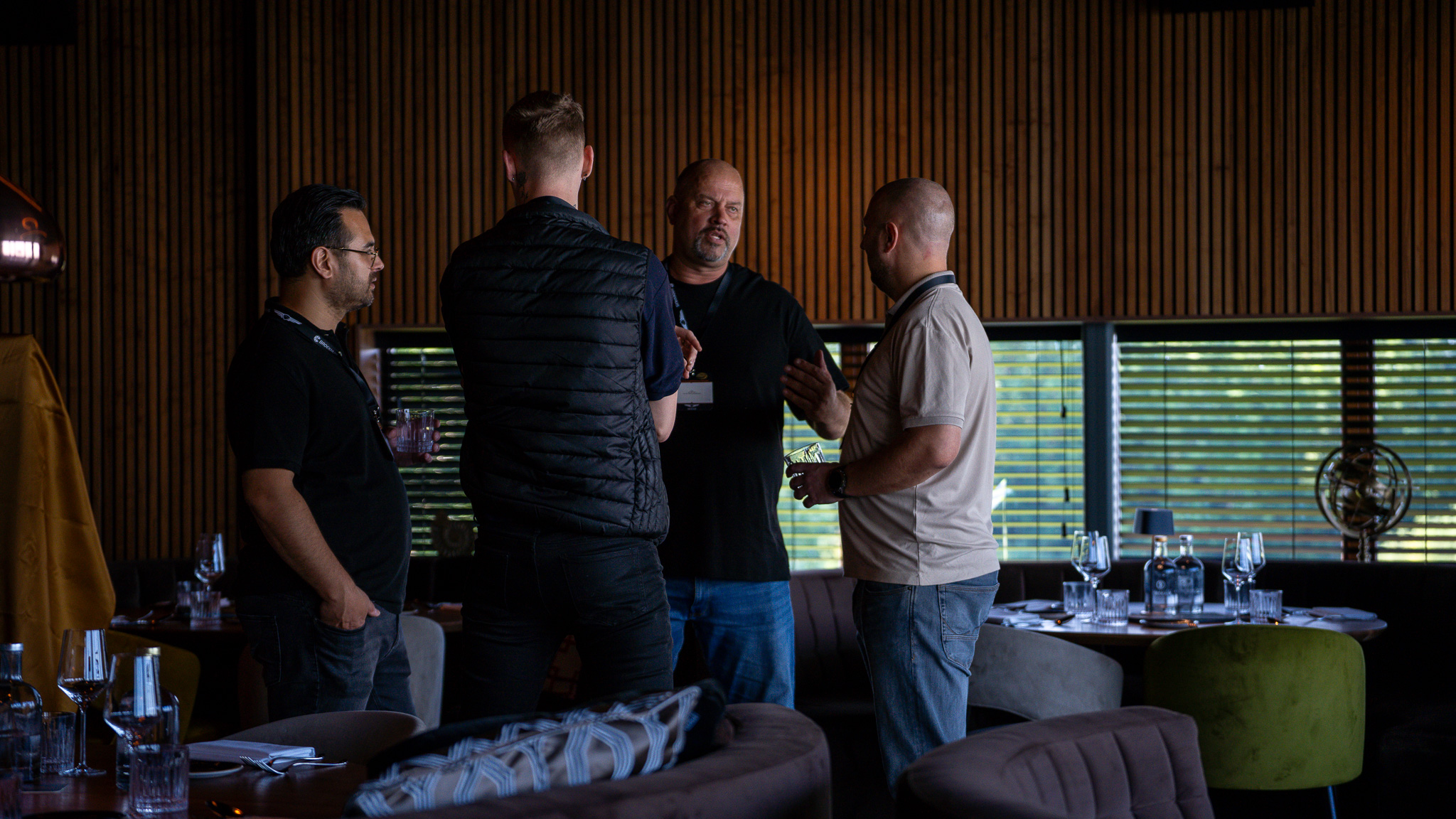 Vier personen in gesprek in een warm verlichte restaurantsetting met gedekte tafels en houten wandpanelen.