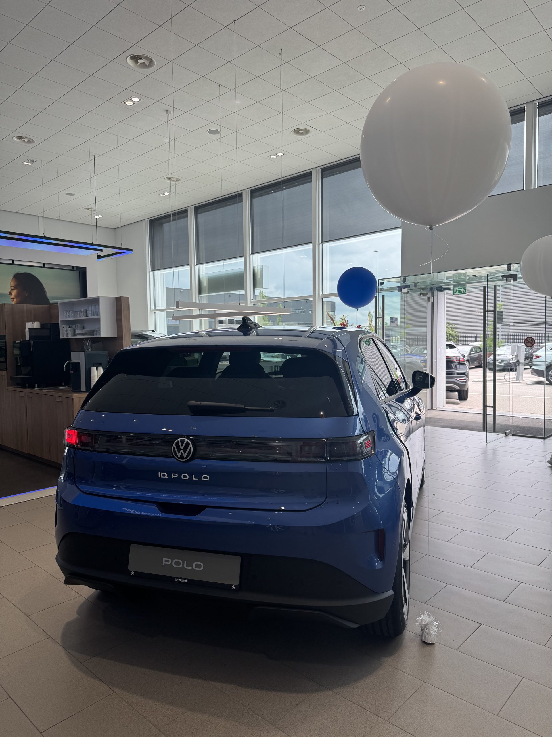 Blauwe Volkswagen Polo in moderne showroom met grote ramen en feestelijke blauwe en witte ballonnen.
