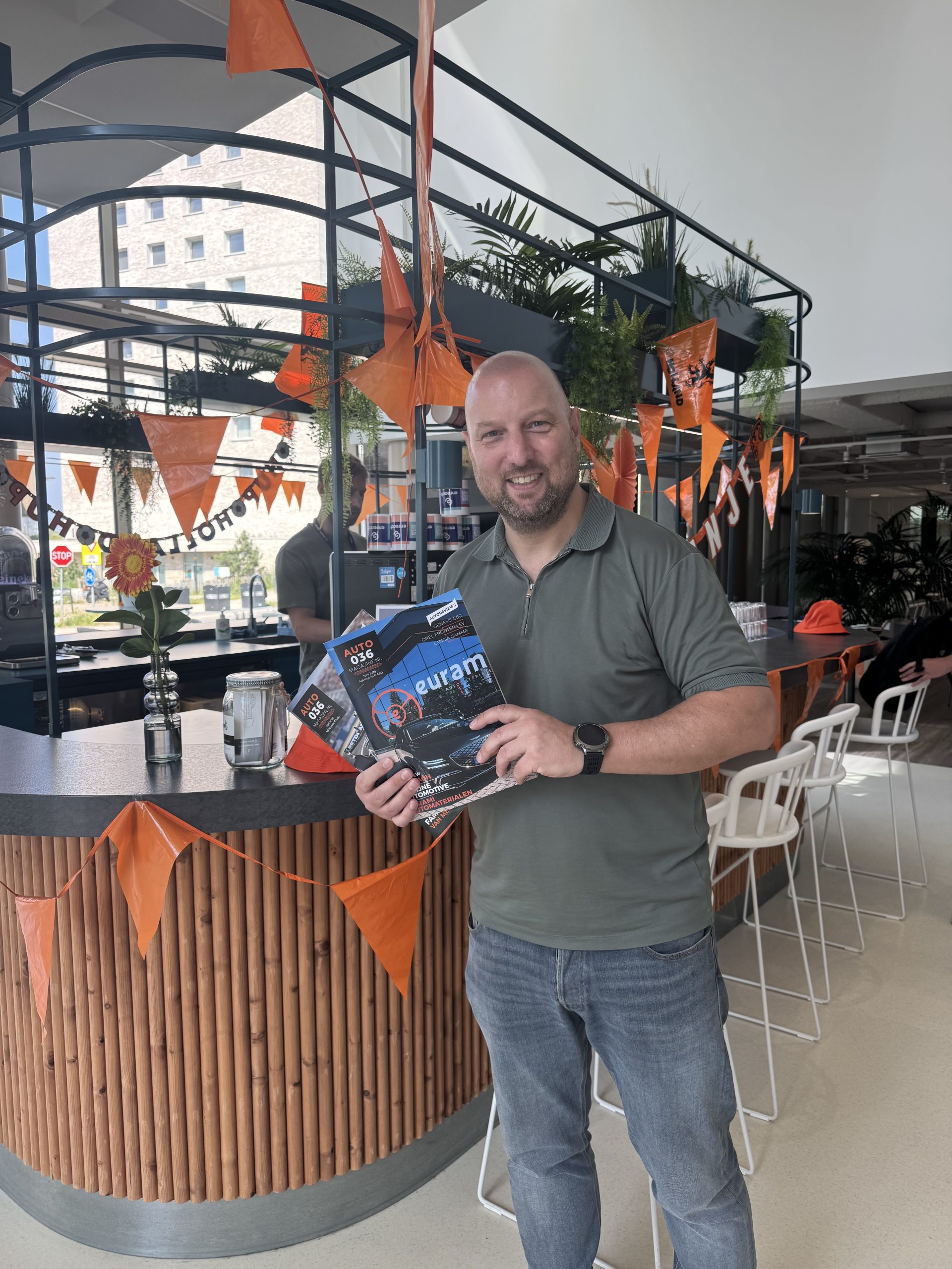 Persoon met Autoweek‑magazines in een modern interieur met oranje vlaggetjes en planten.