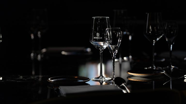 Close‑up van twee Bordeaux‑wijnglazen op een sfeervol gedekte tafel met zacht warm licht.